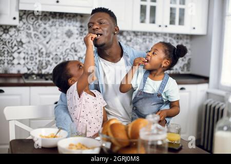 Glücklicher afrikanischer Vater hält niedliche Töchter auf Knien, während sie ihn mit hausgemachten Keksen füttern. Hübscher Mann und zwei Mädchen, die beim Frühstück in der modernen Küche Spaß haben. Stockfoto