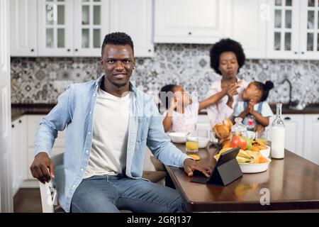 afroamerikanischer Mann, der auf einer hellen Küche mit digitalem Tablet sitzt und auf die Kamera schaut. Verwischen Sie den Hintergrund einer charmanten Frau, die während des Frühstücks mit Töchtern spielt. Schöne Familie zu Hause. Stockfoto