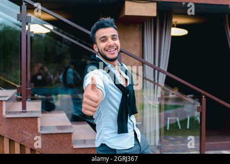 Fröhlicher junger Mann sieht muslimisch lächelnd und zeigt Handgesten Cool , ist neben dem Restaurant auf dem Hintergrund der Treppe im Freien. Stockfoto