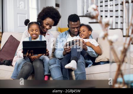 Fröhliche afroamerikanische vierköpfige Familie mit Joysticks und digitalem Tablet zum Spielen von Videospielen auf einer bequemen Couch. Eltern mit zwei Töchtern verbringen Zeit zu Hause mit Spaß. Stockfoto
