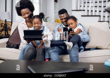 Glückliche afroamerikanische vierköpfige Familie mit Joysticks und digitalem Tablet zum Spielen von Videospielen auf der bequemen Couch. Eltern mit zwei Töchtern verbringen Zeit zu Hause mit Spaß. Stockfoto