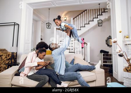 afroamerikanische Mutter schaut sich Cartoons mit ihrer Tochter auf einem digitalen Tablet an, während ihr Vater mit einer anderen Tochter spielt. Glückliche Eltern genießen die Zeit mit ihren Kindern. Stockfoto