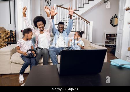 Fröhliche afroamerikanische Eltern und ihre zwei niedlichen Töchter spielen Videospiele auf dem Laptop, während sie auf der bequemen Couch sitzen. Positive Emotionen vom Sieg. Freizeit zu Hause. Stockfoto