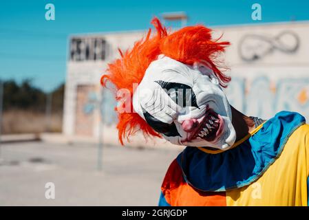 Nahaufnahme eines gruseligen bösen Clowns mit roten Haaren und einem bunten Kostüm, der Gesichter macht, während er den Betrachter an einem einsamen Ort im Freien anstarrt Stockfoto