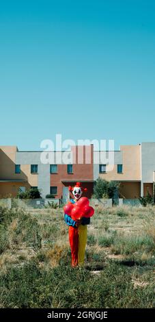 Ein gruseliger Clown, der ein farbenfrohes Kostüm trägt, einen Haufen roter Ballons in der Hand hält, auf einem leeren Grundstück steht, in einem vertikalen Format, das für den Mob verwendet werden kann Stockfoto