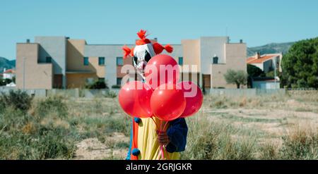 Nahaufnahme eines gruseligen Clowns, der ein farbenprächtiges Gelb-, Rot- und Blaukostüm trägt, einen Haufen roter Luftballons in der Hand hält, auf einem leeren Grundstück steht, in der Hand Stockfoto