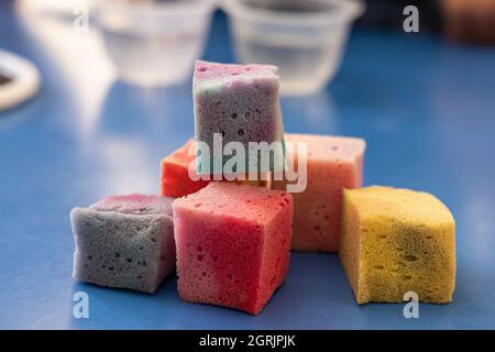 Ein in quadratische Würfel geschnittener Schwamm, in verschiedene Farben getaucht, auf einen blauen Tisch zum Trocknen gelegt, neben den mit Wasser gefüllten Kristallschüsseln. Stockfoto