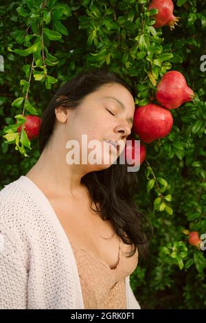 Porträt einer jungen Frau mit geschlossenen Augen posiert mit reifen roten Granatäpfel Baum. Konzept der gesunden Herbst und Winter Lebensstil und Ernährung, im Freien wit Stockfoto