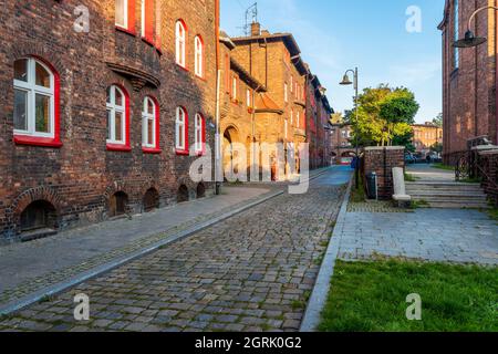 Blick auf die schmale, kopfsteingepflasterte Straße im traditionellen, schlesischen Stadtteil Nikiszowiec in Katowice, Polen. Alte Backsteinhäuser mit rot gestrichenen Fensterrahmen Stockfoto