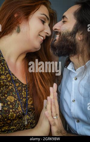 Porträt eines jungen Paares, in dem eine lange rothaarige Frau und ein bärtiger Mann mit langen Haaren einander mit gefalteten Händen in die Augen schauen Stockfoto