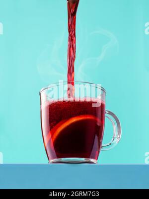 Gießen Sie heißen Wein in einem Glas Becher auf einem blauen cyanfarbenen Hintergrund. Weihnachtlicher Glühwein in einem Becher, isoliert auf einem farbigen Hintergrund. Stockfoto