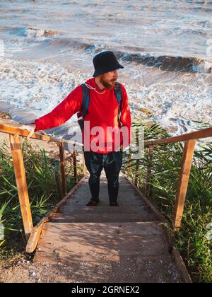 Bärtiger Tausendjähriger Mann in rotem Kapuzenpullover mit Rucksack Herbst Meer Hintergrund trockene Schilf authentische männliche Tourist Lifestyle Foto Stockfoto