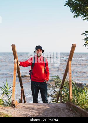 Bärtiger Tausendjähriger Mann in rotem Kapuzenpullover mit Rucksack Herbst Meer Hintergrund trockene Schilf authentische männliche Tourist Lifestyle Foto Stockfoto