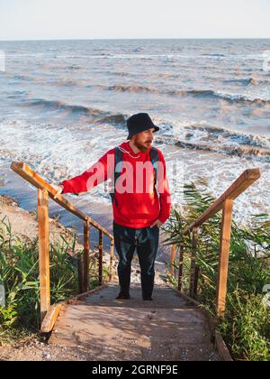 Bärtiger Tausendjähriger Mann in rotem Kapuzenpullover mit Rucksack Herbst Meer Hintergrund trockene Schilf authentische männliche Tourist Lifestyle Foto Stockfoto