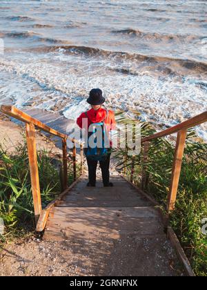 Bärtiger Tausendjähriger Mann in rotem Kapuzenpullover mit Rucksack Herbst Meer Hintergrund trockene Schilf authentische männliche Tourist Lifestyle Foto Stockfoto