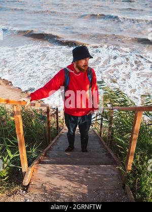 Bärtiger Tausendjähriger Mann in rotem Kapuzenpullover mit Rucksack Herbst Meer Hintergrund trockene Schilf authentische männliche Tourist Lifestyle Foto Stockfoto