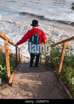 Bärtiger Tausendjähriger Mann in rotem Kapuzenpullover mit Rucksack Herbst Meer Hintergrund trockene Schilf authentische männliche Tourist Lifestyle Foto Stockfoto