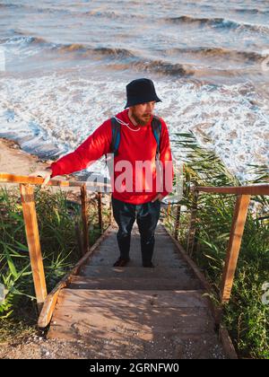 Bärtiger Tausendjähriger Mann in rotem Kapuzenpullover mit Rucksack Herbst Meer Hintergrund trockene Schilf authentische männliche Tourist Lifestyle Foto Stockfoto