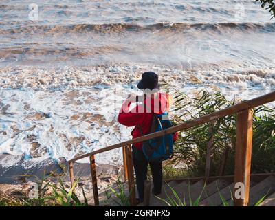 Bärtiger Tausendjähriger Mann in rotem Kapuzenpullover mit Rucksack Herbst Meer Hintergrund trockene Schilf authentische männliche Tourist Lifestyle Foto Stockfoto
