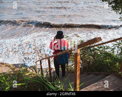 Bärtiger Tausendjähriger Mann in rotem Kapuzenpullover mit Rucksack Herbst Meer Hintergrund trockene Schilf authentische männliche Tourist Lifestyle Foto Stockfoto