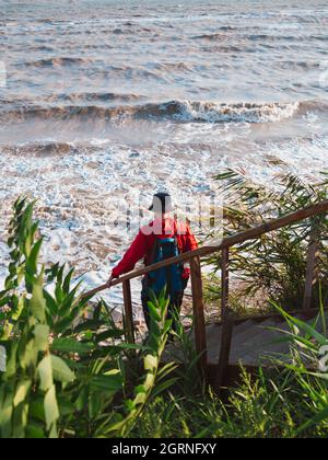 Bärtiger Tausendjähriger Mann in rotem Kapuzenpullover mit Rucksack Herbst Meer Hintergrund trockene Schilf authentische männliche Tourist Lifestyle Foto Stockfoto