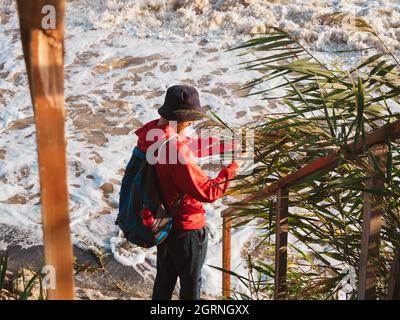Bärtiger Tausendjähriger Mann in rotem Kapuzenpullover mit Rucksack Herbst Meer Hintergrund trockene Schilf authentische männliche Tourist Lifestyle Foto Stockfoto