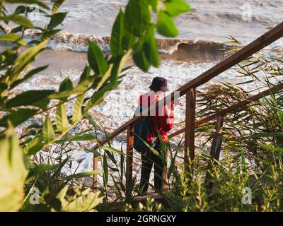 Bärtiger Tausendjähriger Mann in rotem Kapuzenpullover mit Rucksack Herbst Meer Hintergrund trockene Schilf authentische männliche Tourist Lifestyle Foto Stockfoto