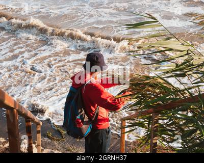 Bärtiger Tausendjähriger Mann in rotem Kapuzenpullover mit Rucksack Herbst Meer Hintergrund trockene Schilf authentische männliche Tourist Lifestyle Foto Stockfoto