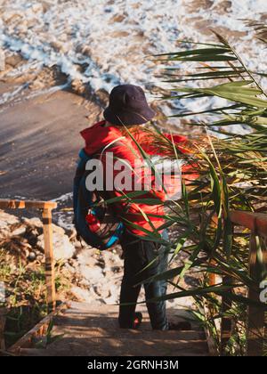 Bärtiger Tausendjähriger Mann in rotem Kapuzenpullover mit Rucksack Herbst Meer Hintergrund trockene Schilf authentische männliche Tourist Lifestyle Foto Stockfoto