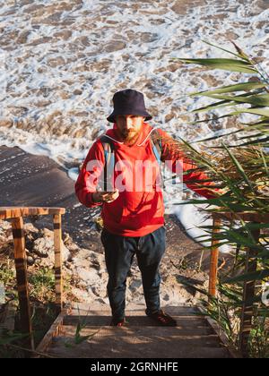 Bärtiger Tausendjähriger Mann in rotem Kapuzenpullover mit Rucksack Herbst Meer Hintergrund trockene Schilf authentische männliche Tourist Lifestyle Foto Stockfoto