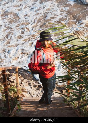 Bärtiger Tausendjähriger Mann in rotem Kapuzenpullover mit Rucksack Herbst Meer Hintergrund trockene Schilf authentische männliche Tourist Lifestyle Foto Stockfoto