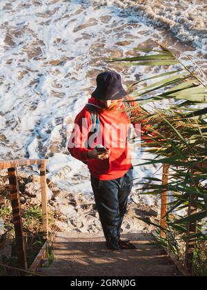 Bärtiger Tausendjähriger Mann in rotem Kapuzenpullover mit Rucksack Herbst Meer Hintergrund trockene Schilf authentische männliche Tourist Lifestyle Foto Stockfoto