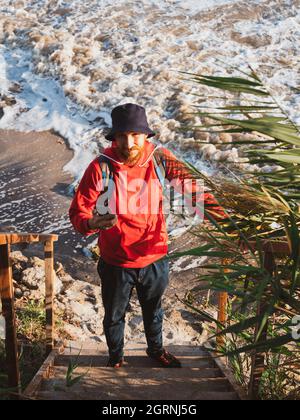 Bärtiger Tausendjähriger Mann in rotem Kapuzenpullover mit Rucksack Herbst Meer Hintergrund trockene Schilf authentische männliche Tourist Lifestyle Foto Stockfoto