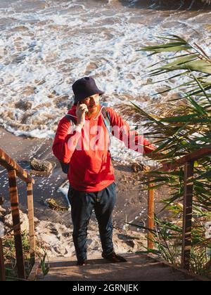 Bärtiger Tausendjähriger Mann in rotem Kapuzenpullover mit Rucksack Herbst Meer Hintergrund trockene Schilf authentische männliche Tourist Lifestyle Foto Stockfoto