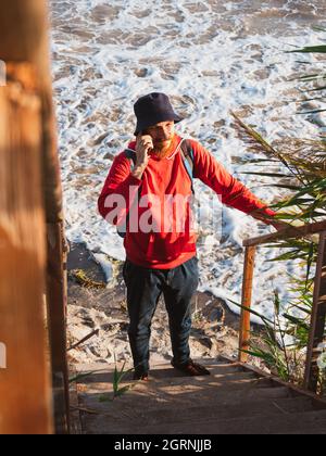 Bärtiger Tausendjähriger Mann in rotem Kapuzenpullover mit Rucksack Herbst Meer Hintergrund trockene Schilf authentische männliche Tourist Lifestyle Foto Stockfoto