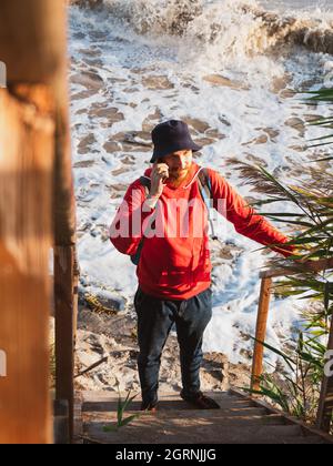 Bärtiger Tausendjähriger Mann in rotem Kapuzenpullover mit Rucksack Herbst Meer Hintergrund trockene Schilf authentische männliche Tourist Lifestyle Foto Stockfoto