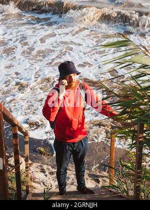 Bärtiger Tausendjähriger Mann in rotem Kapuzenpullover mit Rucksack Herbst Meer Hintergrund trockene Schilf authentische männliche Tourist Lifestyle Foto Stockfoto