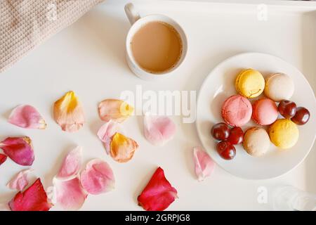 Tasse Kaffee mit Milch, Teller Makronen und Kirschen auf weißem Tisch. Überraschung zum Valentinstag. Köstliches romantisches Frühstück mit Rosenblättern. Morgengetränk mit Dessert. Stockfoto