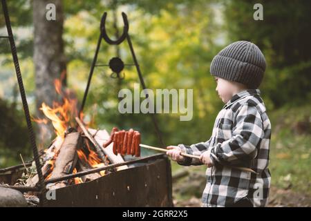 Ein kleines Kind in einem karierten Hemd und einem grauen Hut fritiert Würstchen am Lagerfeuer im Wald. Kindheit mit naturliebenden Konzept Stockfoto
