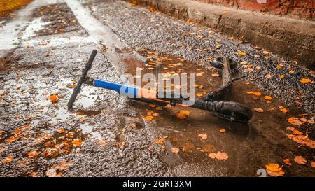 Elektroroller liegt auf dem Asphalt in einer Pfütze mit gelben Herbstblättern. Persönliche Transportprobleme, Verkehrsunfälle, Mitfahren und saisonal Stockfoto