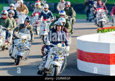 Quadrophenia-Star Phil Daniels beim Goodwood Revival 2014 führt eine Gruppe von „Mods“ auf der Rennstrecke mit einem Motorroller in Retro-Kleidung durch Stockfoto