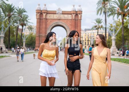 Junge multirassische Freundinnen in modischer Kleidung reden und lachen beim Bummeln auf dem Platz von Barcelona City mit Triumphbogen im Hintergrund am Sommertag Stockfoto