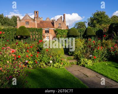 Chenies Herrenhaus im Herbst vom versunkenen Garten aus gesehen mit vielen Dahlien, die noch in Blüte stehen. Farbenfrohe Pflanzenränder, beschnittene Eibenbäume und Spaliere. Stockfoto