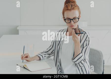 Junge konzentrierte Ingwer-Frau freiberuflich mit Spracherkennungssystem auf dem Smartphone Stockfoto