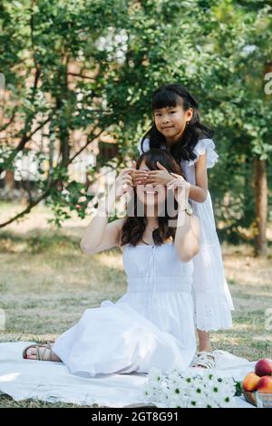 Das kleine Mädchen hat Spaß mit ihrer Mutter und bedeckt ihre Augen mit Handflächen. Beide trugen weiße Kleider und picknicken in einem Park, genießend Stockfoto
