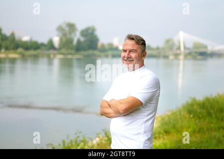 Ein älterer Mann, der an einem sonnigen Frühlingstag mit gekreuzten Armen am Ufer eines Flusses steht und mit einem Lächeln über das Wasser blickt Stockfoto