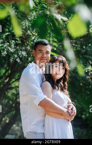 Schönes Porträt eines asiatischen Paares in weißer Kleidung in einem Park. Mann hält Frau von hinten. Sie genießen die letzten warmen Tage des frühen Herbstes. Stockfoto