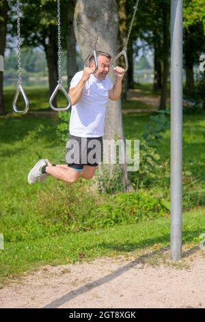 Ein älterer Mann, der in einem Park in einer Sportanlage im Freien an Übungsringen in einem Gesundheits-, Fitness- und aktiven Lifestyle-Konzept arbeitet Stockfoto