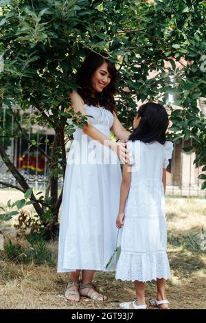 Mutter und Tochter stehen unter einem kurzen Baum in einem Park und blicken einander mit Liebe und Anbetung an. Beide tragen weiße Kleider und genießen la Stockfoto