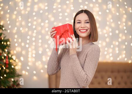 Porträt von jungen glücklichen Frau roten Lippen Blick auf verpackte Geschenkbox. Nahaufnahme Zufriedene Frau erhielt Geschenk-Box. Festliche Weihnachten Lichter Hintergrund. Stockfoto